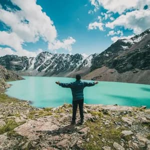 Eine Person steht mit ausgestreckten Armen auf einem Felsvorsprung und blickt über einen türkisen Alpensee, der von schneebedeckten Bergen umgeben ist.