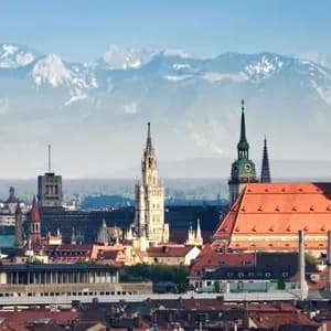 Un paesaggio urbano panoramico con tetti rossi e campanili, e una grande catena montuosa innevata sullo sfondo.