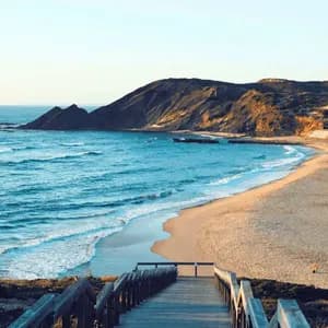 Vista da una scala di legno che si affaccia su una spiaggia sabbiosa con onde blu dell'oceano e una grande scogliera rocciosa.