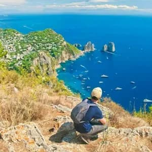 Una persona con uno zaino si accovaccia su una scogliera rocciosa, guardando una città costiera e un mare blu punteggiato di barche.