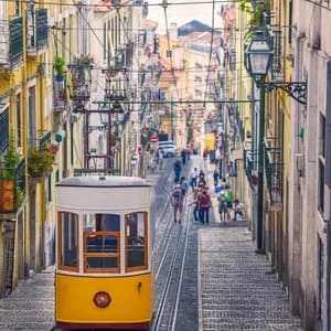 A yellow funicular tram travels up a steep, narrow cobblestone street lined with colorful buildings, with a view of the water in the distance.