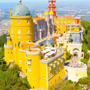 Una vista aerea di un grande castello colorato con torri gialle e rosse, situato su una collina verde e alberata sotto un cielo azzurro e limpido.