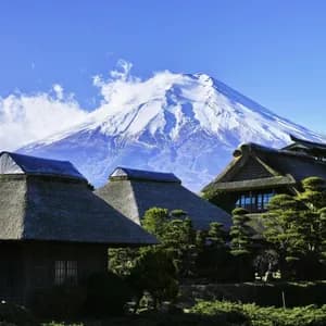 Case tradizionali con tetto in paglia e una ruota idraulica si ergono di fronte a una grande montagna innevata, sotto un cielo azzurro.