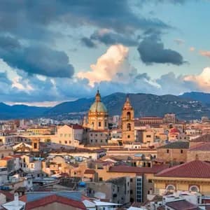 Paesaggio urbano con edifici storici, una cupola e un campanile prominenti, sullo sfondo di montagne sotto un cielo nuvoloso.