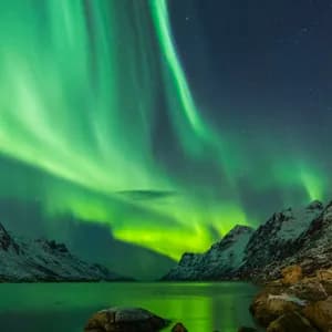 L'Aurora Boreale risplende verde in un cielo stellato sopra un fiordo circondato da montagne innevate.
