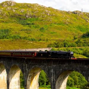 Ein schwarzer Dampfzug mit roten Waggons fährt über ein steinernes Viadukt, mit einem üppig grünen Berg im Hintergrund.