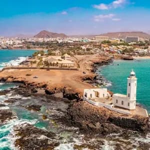 An aerial view of a white lighthouse on a rocky peninsula, with a coastal city and mountains in the background under a blue sky.