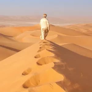 Una persona in abiti tradizionali cammina lungo la cresta di una grande duna di sabbia nel deserto, lasciandosi dietro delle impronte.
