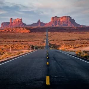 Una lunga strada diritta si estende attraverso un paesaggio desertico verso formazioni rocciose rosse all'orizzonte sotto un tramonto tenue.
