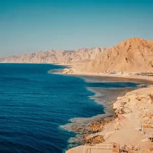 A panoramic view of a deep blue sea meeting a rugged, arid coastline with mountains under a clear sky.