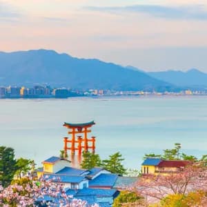 Un torii rosso si erge nel mare, con ciliegi in fiore e case tradizionali in primo piano e montagne in lontananza.