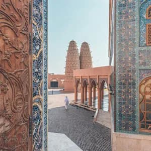 Una persona in abiti bianchi tradizionali cammina in un cortile con torri colombare, vista tra una porta intagliata e un muro a mosaico.