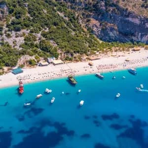 An aerial view of a crowded white sand beach at the base of a green hill, with many boats floating in the turquoise sea.