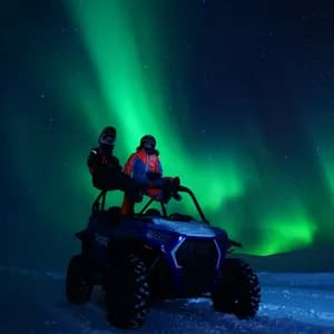 Dos personas con cascos sentadas en un vehículo todoterreno en la nieve, observando la aurora boreal iluminar el cielo nocturno estrellado.