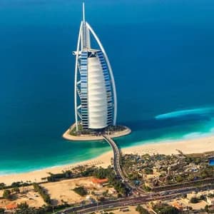 Veduta aerea di un hotel a forma di vela su una piccola isola artificiale, collegata da un ponte a una costa sabbiosa e a una città.