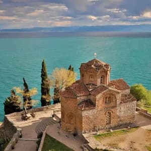 Una storica chiesa in pietra con il tetto di tegole rosse si erge su una scogliera che domina un vasto specchio d'acqua turchese sotto un cielo nuvoloso.