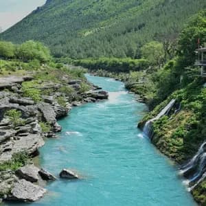 Une rivière turquoise traverse un canyon rocheux, avec de petites cascades dévalant une colline luxuriante à côté d'un bâtiment.