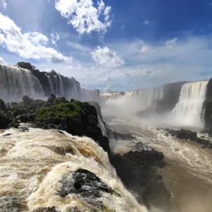 Un'ampia vista di un grande sistema di cascate che si gettano in un canyon nebbioso, con rapide in primo piano sotto un cielo parzialmente nuvoloso