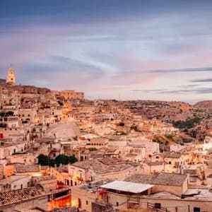 Un borgo di antichi edifici in pietra e un campanile incastonato nella collina, illuminato al crepuscolo accanto a un burrone.