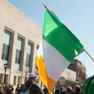 Gros plan sur un drapeau irlandais flottant au soleil lors d'une parade de rue, avec des bâtiments de la ville en arrière-plan.
