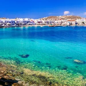 Una cittadina costiera con edifici bianchi si affaccia su una baia di acqua turchese cristallina sotto un cielo azzurro, con una piccola barca sulla riva.