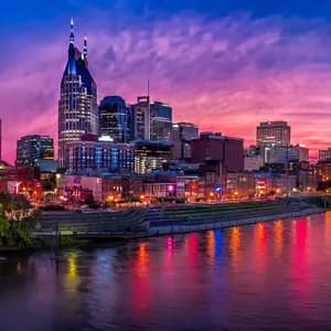 La silhouette illuminée d'une ville se reflète dans une large rivière au coucher du soleil, sous un ciel rose et violet éclatant.