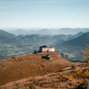 Ein großes Gebäude thront auf einem grasbewachsenen Berggipfel und überblickt ein weites Tal sowie geschichtete Bergketten unter einem klaren Himmel.
