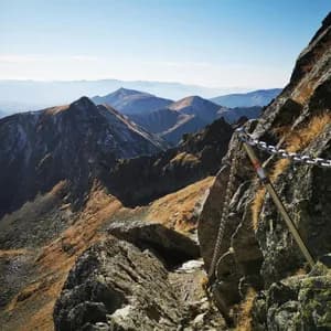 Eine metallene Sicherheitskette verläuft entlang eines schmalen, felsigen Bergpfades mit Blick auf eine weite Bergkette unter klarem Himmel.