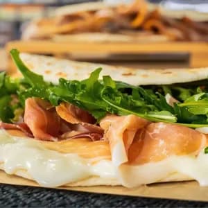 Primo piano di una piadina piegata ripiena di prosciutto, formaggio fuso e rucola fresca su carta marrone.