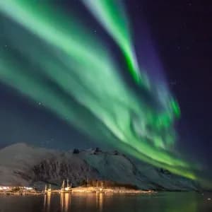 Las auroras boreales verdes surcan un cielo nocturno estrellado sobre montañas cubiertas de nieve y un puerto con barcos.