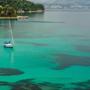 Una barca a vela è ancorata in una baia di acqua cristallina turchese vicino a una costa verde lussureggiante.