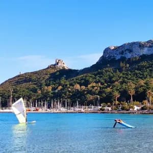 Windsurfistas en aguas tranquilas y azules en una bahía con un puerto deportivo al pie de una montaña verde y rocosa bajo un cielo despejado.