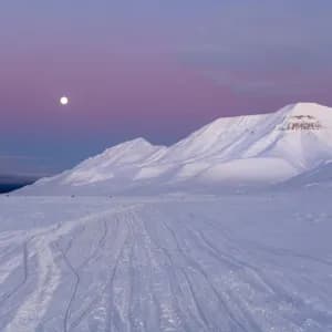 Una luna piena brilla in un cielo crepuscolare violaceo sopra una vasta catena montuosa innevata con tracce nella neve.