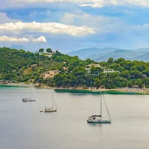 Tre barche a vela galleggiano in una baia tranquilla accanto a una costa verde e collinare con edifici sparsi sotto un cielo nuvoloso.