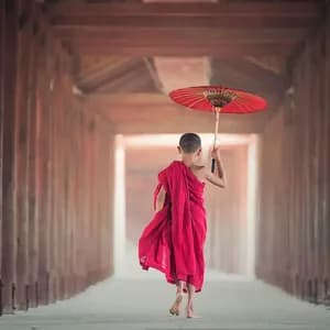 Un giovane monaco a piedi nudi in tuniche rosse cammina lungo un corridoio di legno tenendo un ombrello rosso.