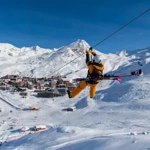 Une personne en équipement de ski descend une tyrolienne avec des skis sur le dos, au-dessus d'un village de montagne enneigé.