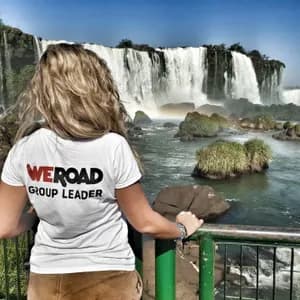 Eine blonde WeRoad Gruppenleiterin steht an einem Geländer und blickt auf einen großen, kaskadierenden Wasserfall unter klarem blauem Himmel.