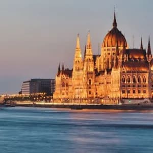 Un edificio gubernamental ornamentado iluminado con luces doradas al anochecer, visto desde el otro lado de un río con barcos.
