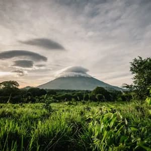 Un volcan avec un nuage lenticulaire au-dessus de son sommet, s'élevant au-dessus d'un paysage verdoyant de champs et de forêt au coucher du soleil.