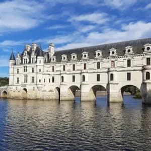 Un château de pierre blanche à tourelles bâti sur des fondations en arc enjambant une rivière, sous un ciel bleu avec quelques nuages.