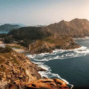 Una vista en picada de una península rocosa y arbolada que incluye un istmo de arena y una pequeña laguna, rodeada por el intenso azul del mar.
