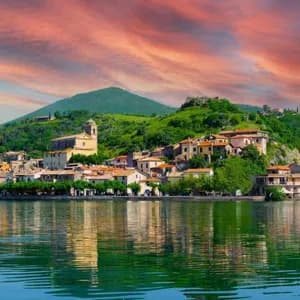 Un borgo lacustre con una chiesa e case dai tetti in terracotta riflesse nell'acqua sotto un cielo al tramonto dai colori vivaci.