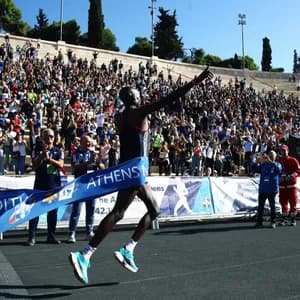 Un corridore taglia il traguardo della maratona di Atene, rompendo il nastro blu mentre la folla esulta dagli spalti dello stadio.