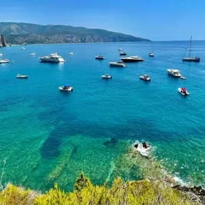 Numerosos barcos y yates están anclados en una bahía con agua clara y turquesa, enmarcada por una costa rocosa y colinas distantes.