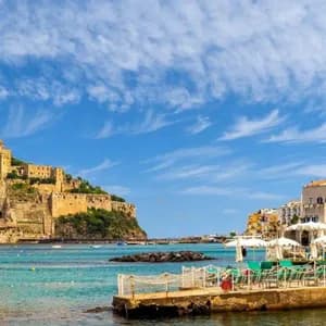 Un castello si erge su una scogliera rocciosa che domina una città costiera e una baia piena di barche sotto un cielo azzurro.