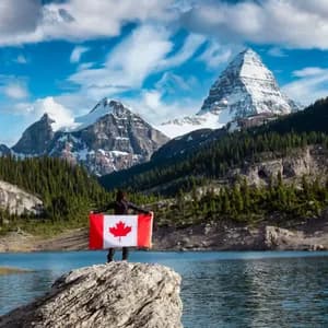 Eine Person hält eine große kanadische Flagge auf einem Felsen mit Blick auf einen Alpensee und schneebedeckte Berge.