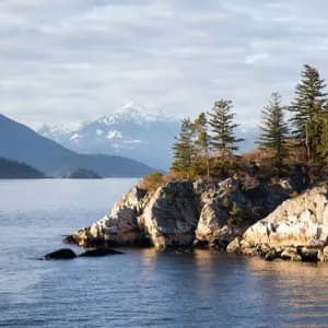 Zwei Personen sitzen auf einer felsigen, bewaldeten Landzunge am Wasser, mit schneebedeckten Bergen, die sich in der Ferne erheben.