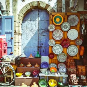 Un puesto de mercado exhibe cerámica colorida, tajines y bolsos de cuero frente a una gran puerta de madera rústica azul.