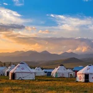 Un accampamento di yurte bianche in un campo erboso con una catena montuosa visibile sullo sfondo sotto un cielo al tramonto.