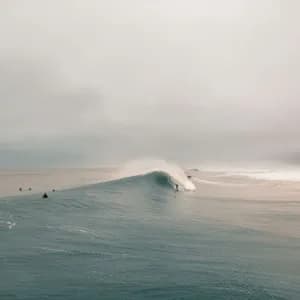 Un surfista cavalca una grande onda tubante nell'oceano, mentre altri surfisti attendono sotto un cielo nuvoloso.